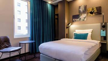 A hotel room with a bed, a chair, a small table, a window, and artwork on the wall.