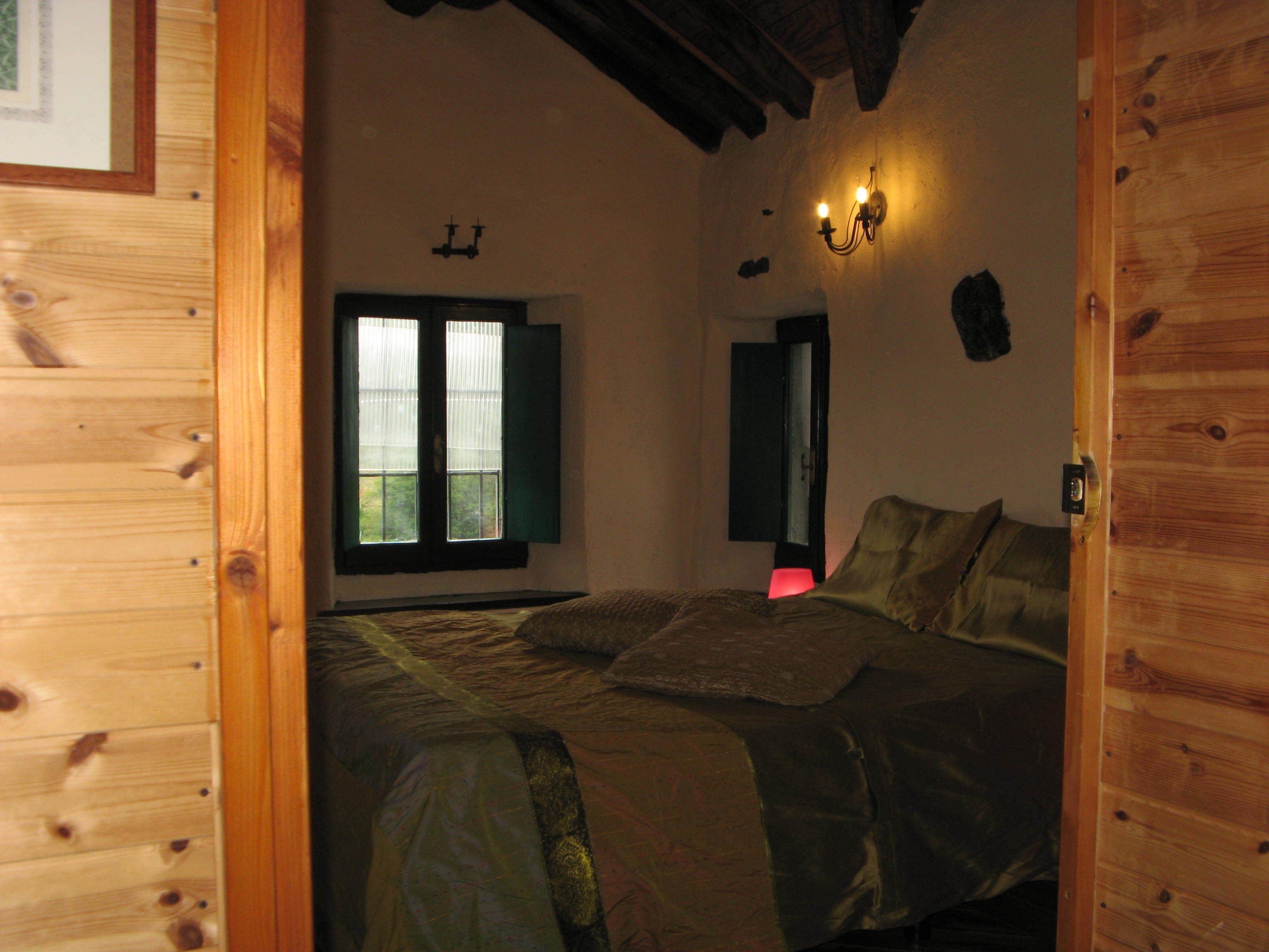 Un dormitorio con puerta de madera, una cama con manta, una mesita de noche, una lámpara de pared y una ventana con contraventanas.
