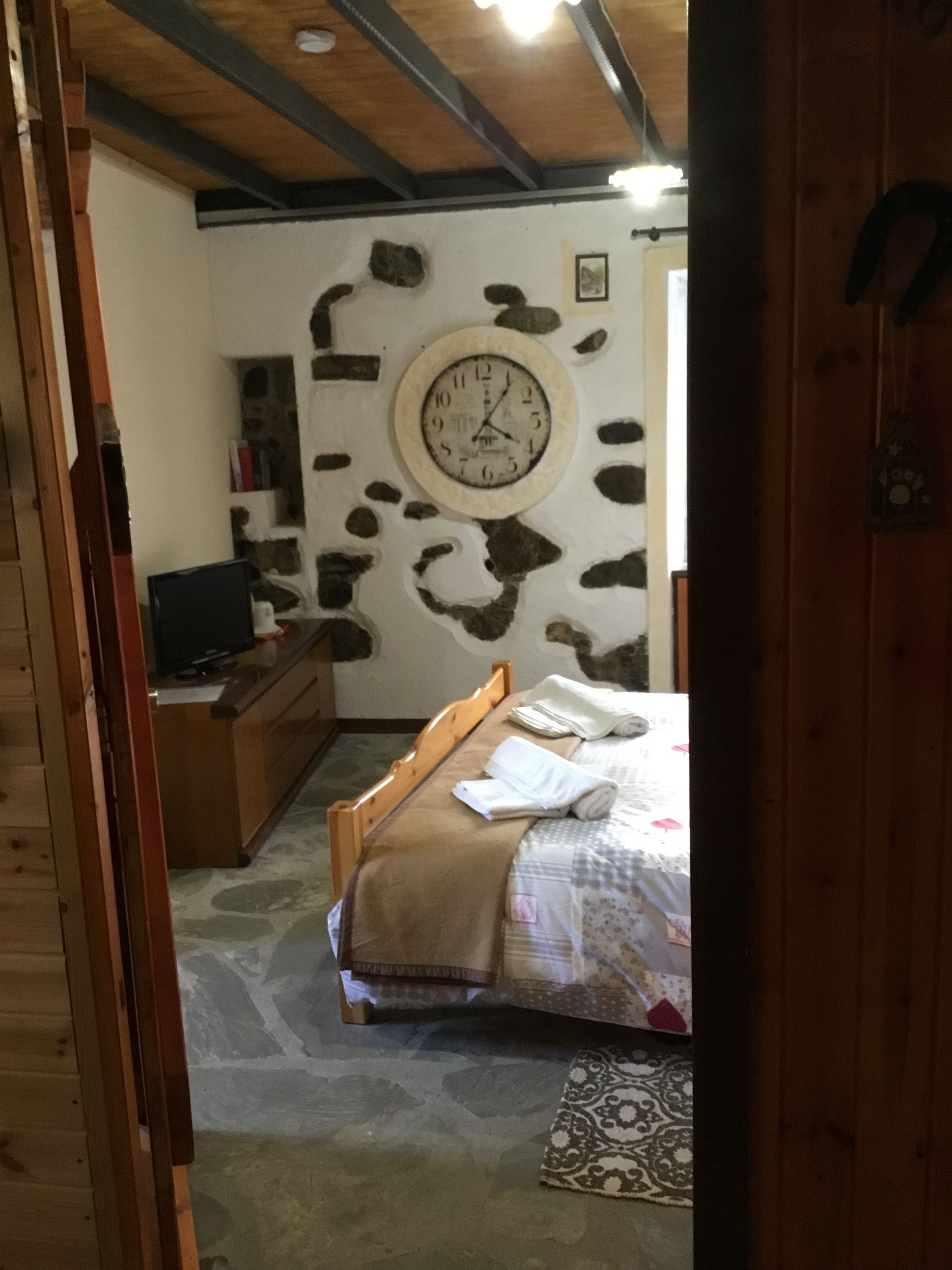 A bedroom with a stone wall, a bed with a patterned bedspread, a wooden headboard, a bedside table, a television, and a clock on the wall.