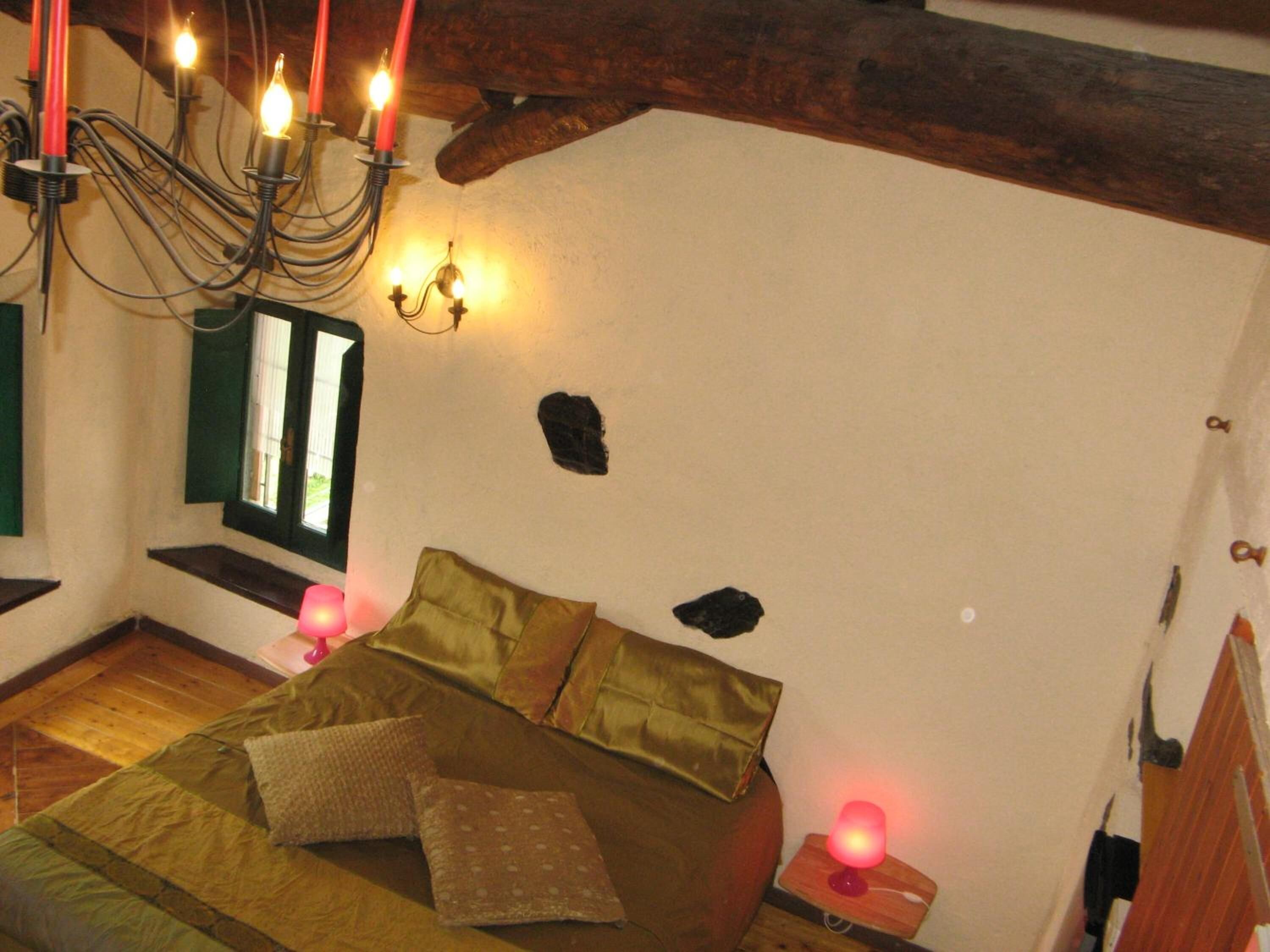 A cozy bedroom with a bed, pillows, a nightstand, a lamp, a window with shutters, and a chandelier.