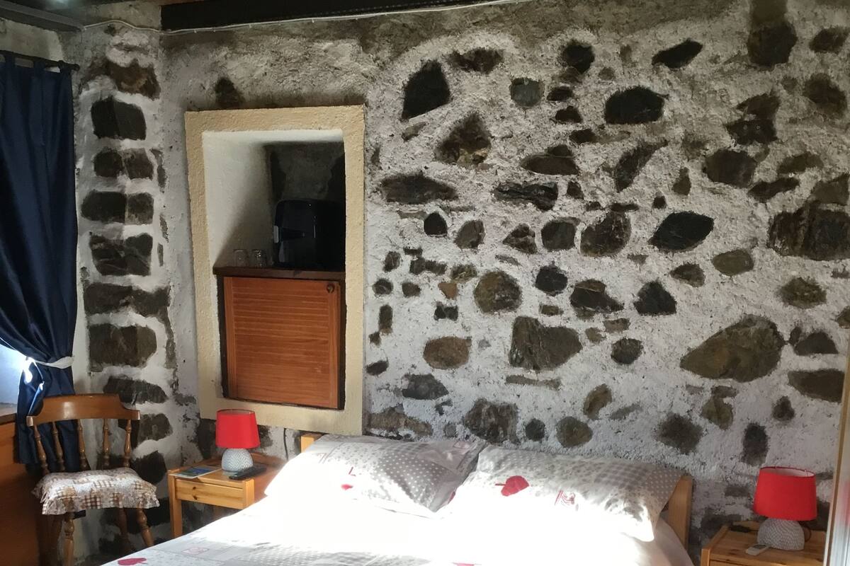Een slaapkamer met een stenen muur, een houten bed, een nachtkastje met lamp en een kleine houten stoel.