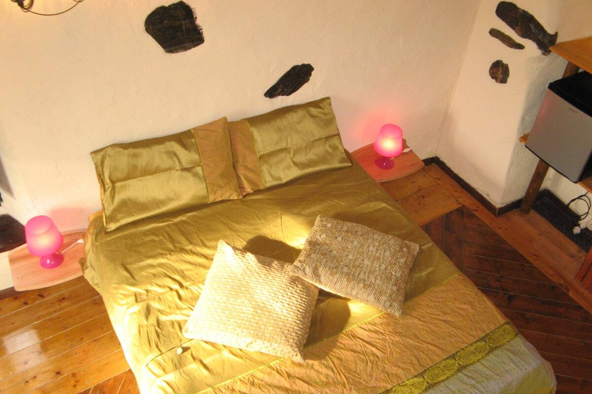 Een bed met een groene plaid, twee kussens en een houten hoofdbord. Er staan twee nachtlampjes met roze kapjes.