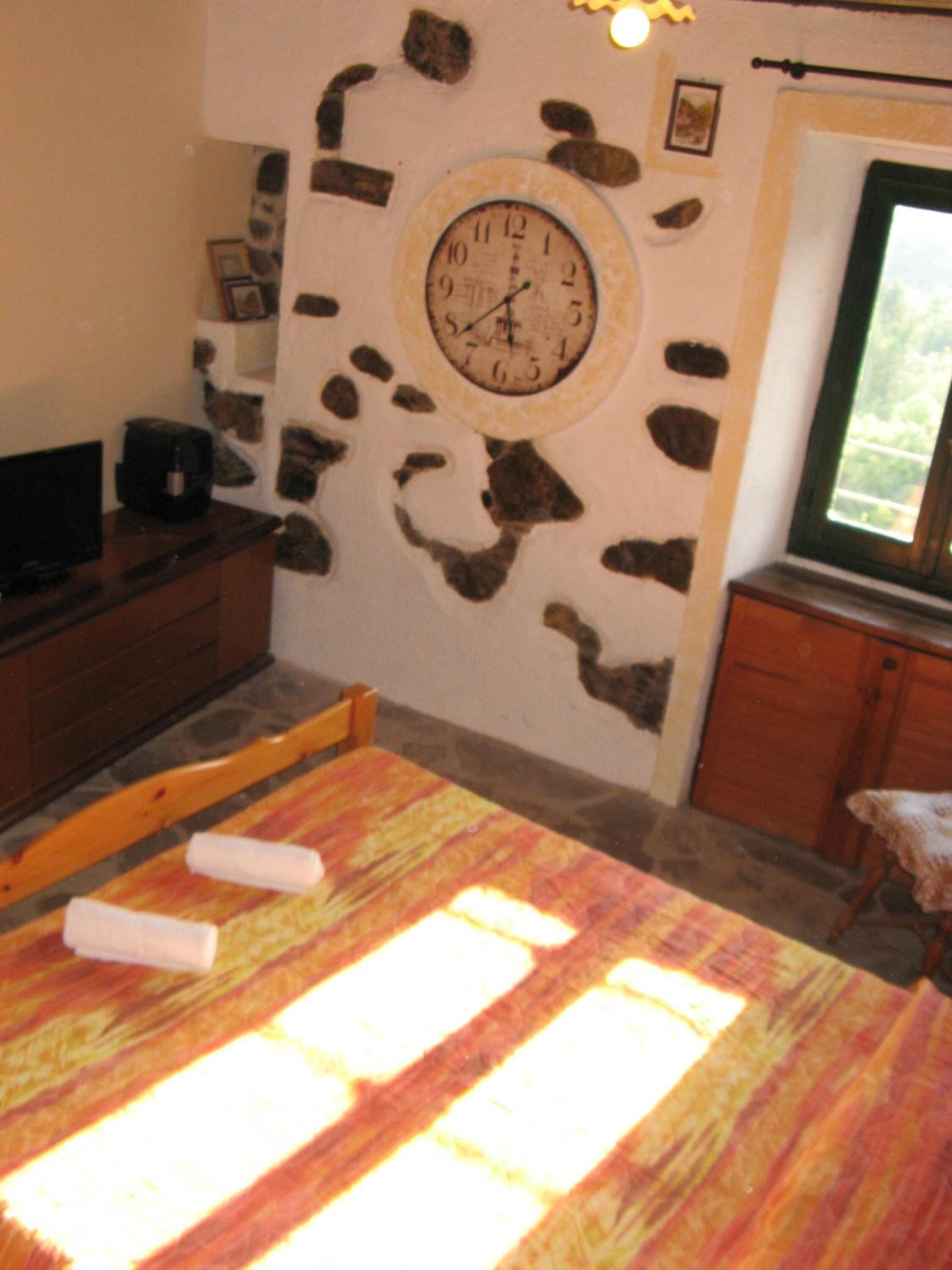 A bedroom with a stone wall, a round clock, a bed with a colorful bedspread, a wooden headboard, a television on a wooden cabinet, and a window with a view of greenery.
