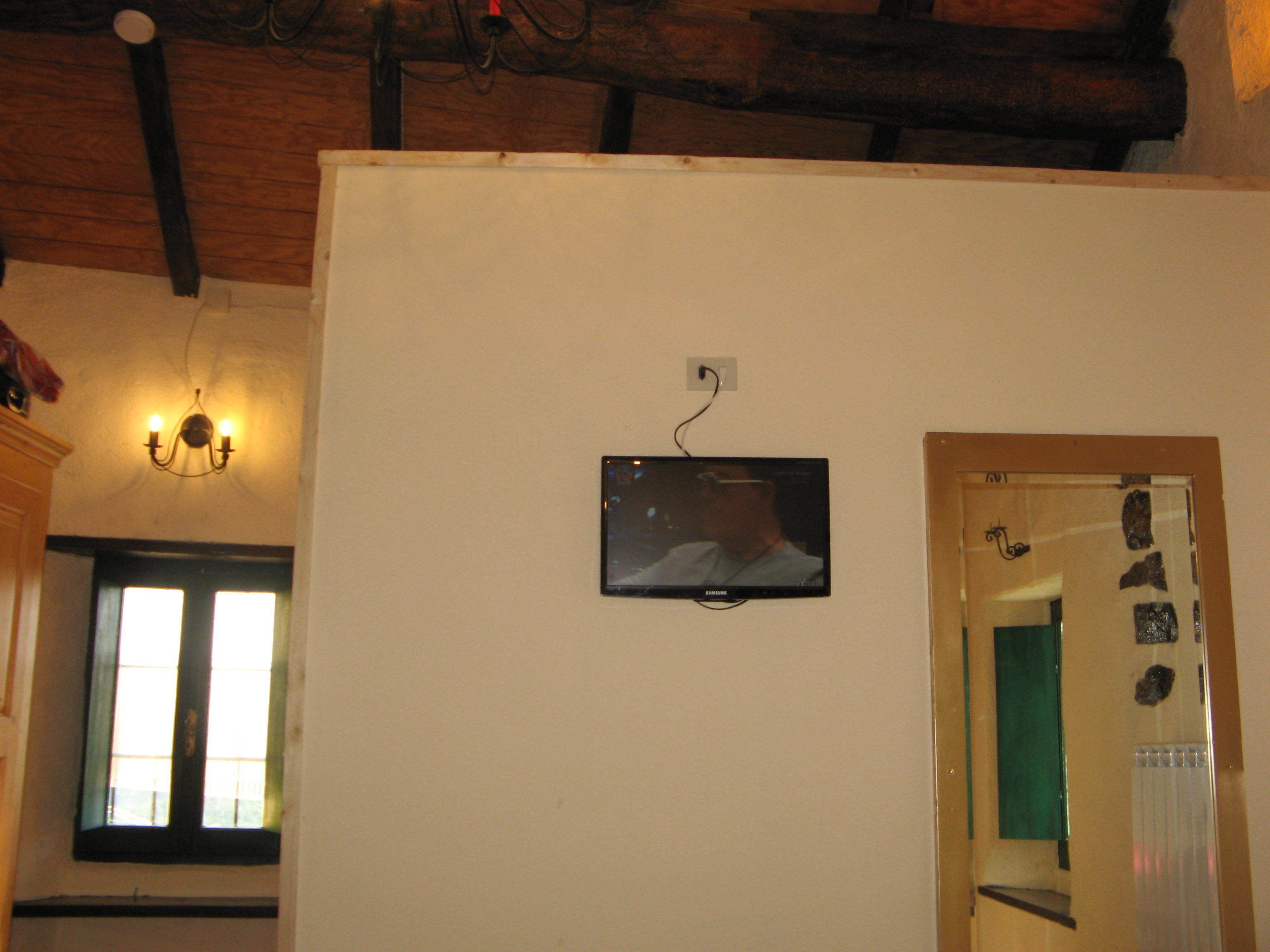 A room with a television mounted on the wall, a window with blinds, and a door with a mirror and decorations.