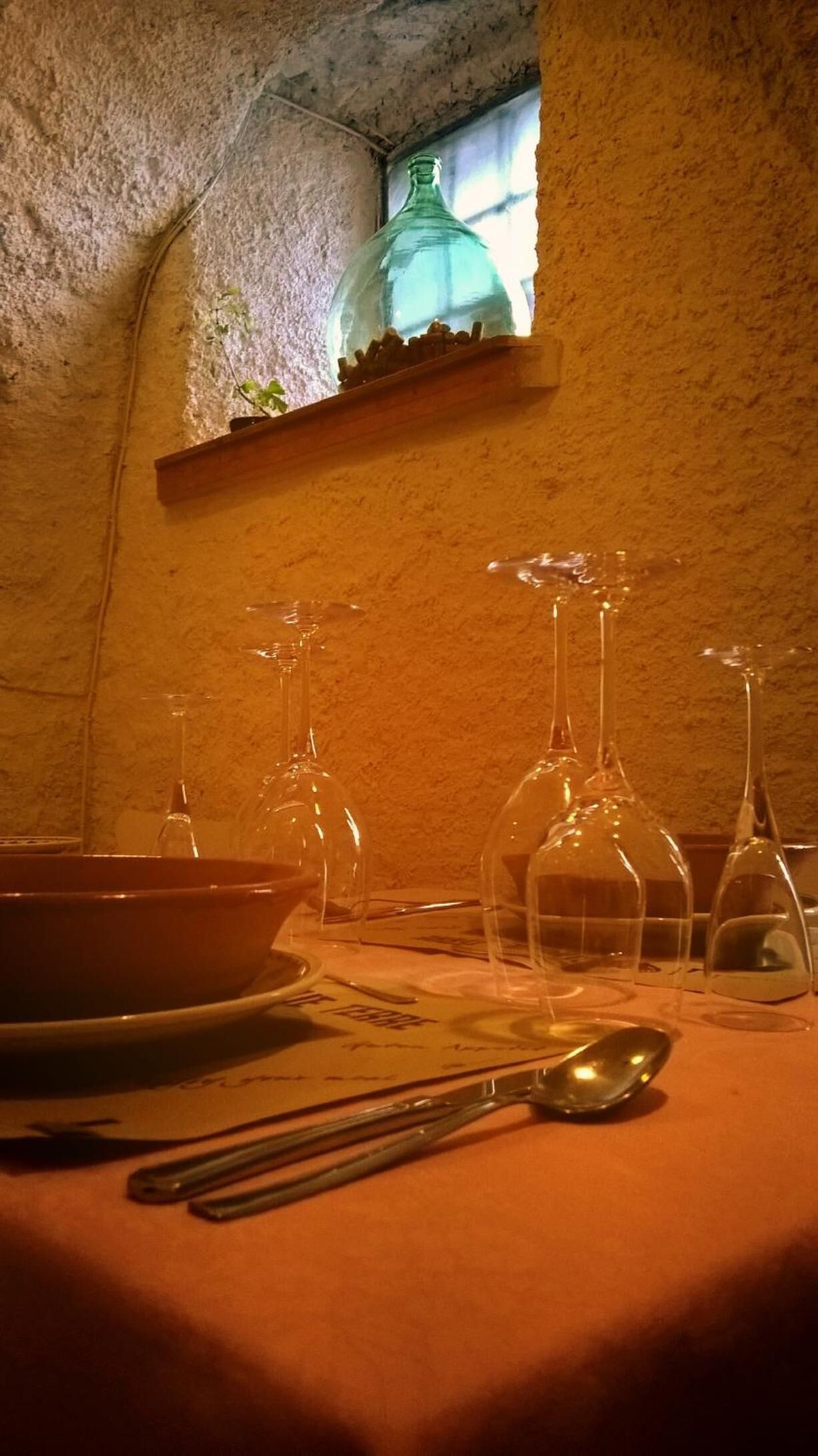 Una mesa dispuesta con copas de vino, un cuenco y cubiertos, con un rústica pared de piedra y una ventana al fondo.