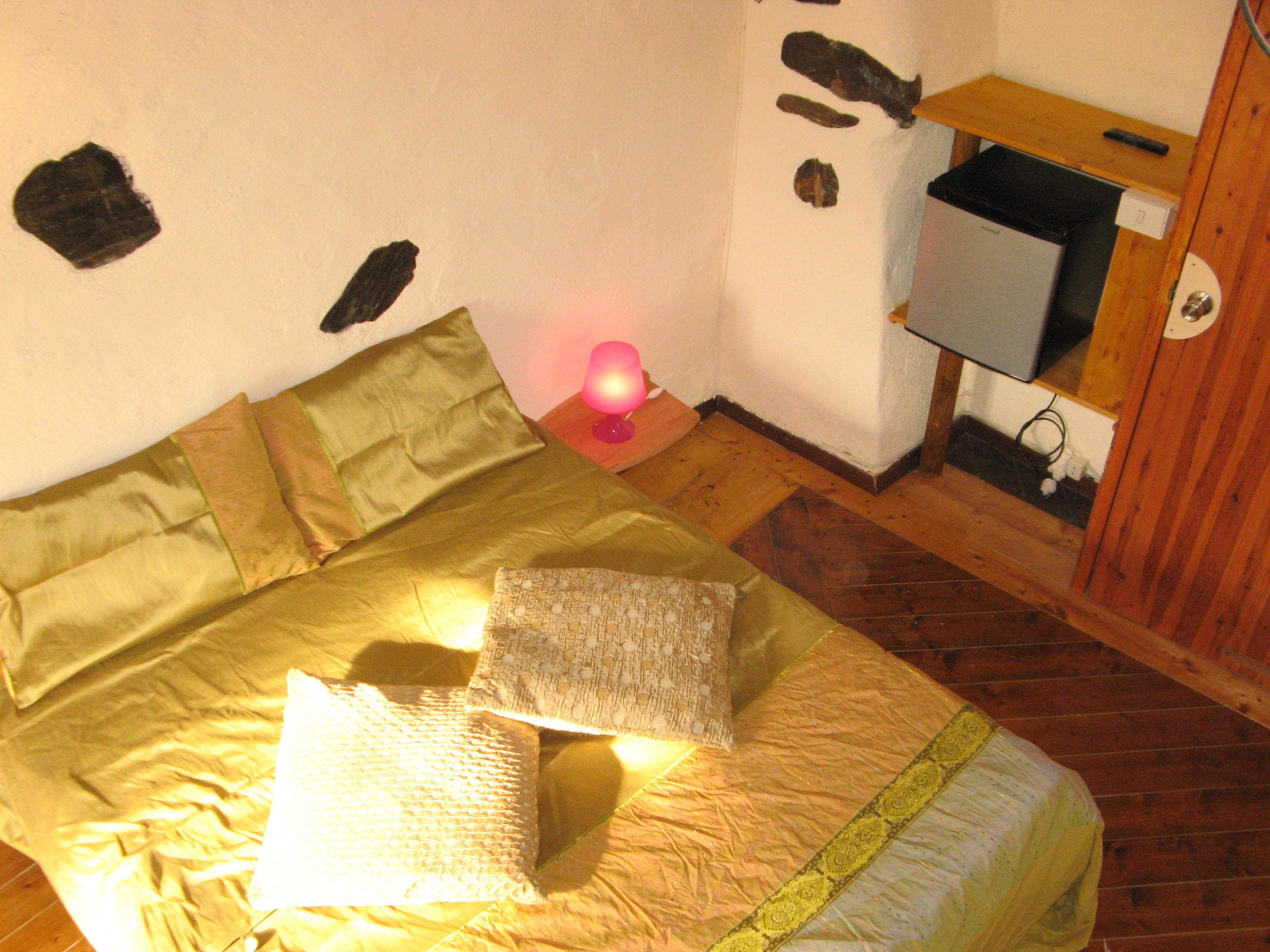 Una cama con colcha y almohadas verdes, una lámpara de noche, un pequeño estante de madera con un mando a distancia y una pared blanca con adornos tipo piedra.