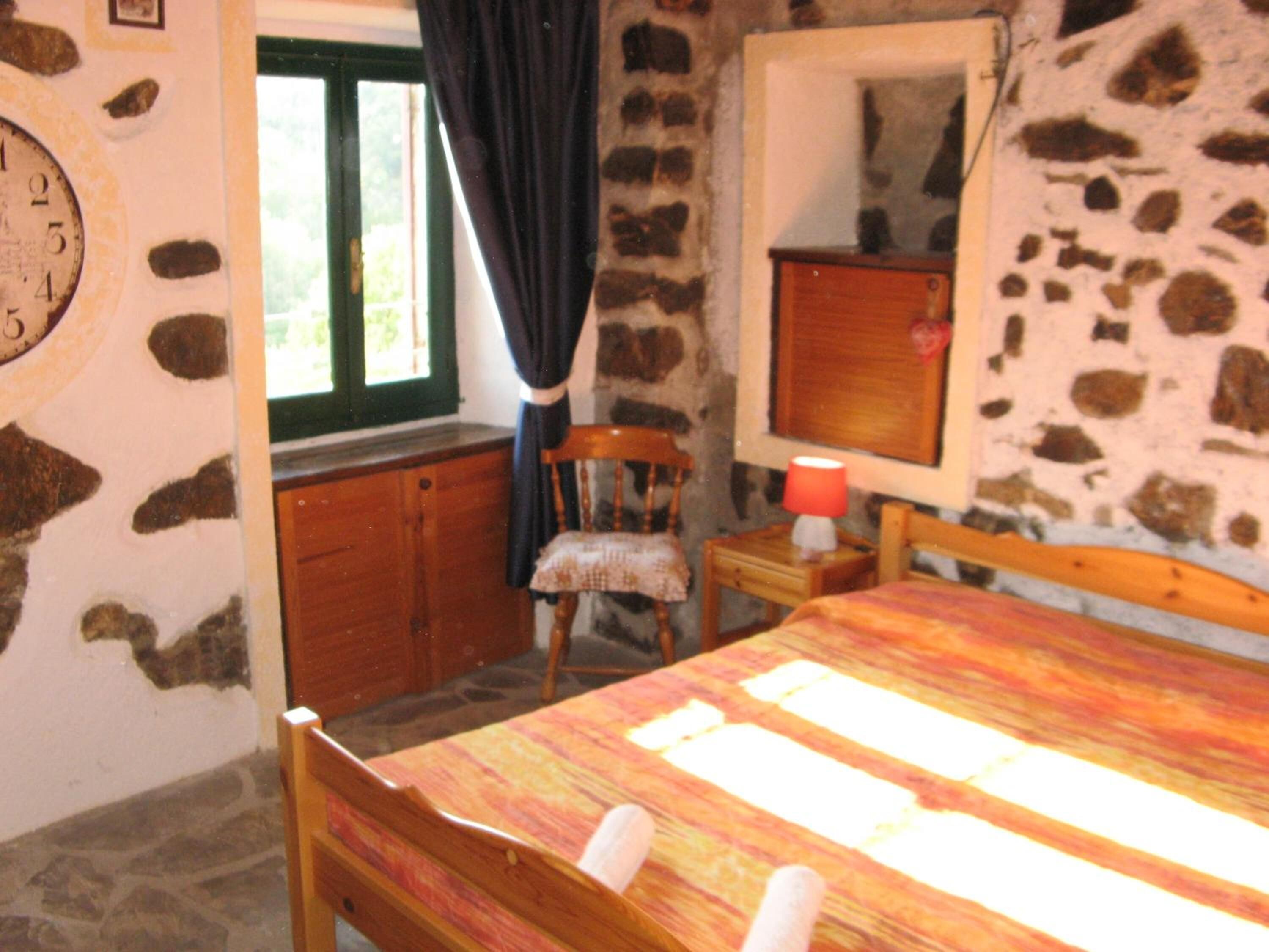 Une chambre avec un mur en pierre, un lit en bois, une chaise, une table de chevet avec une lampe et une fenêtre avec des rideaux.