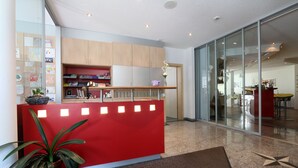 A reception area with a red counter, wooden cabinets, a glass door leading to a dining area, and a potted plant. - Apparthotel Feehof Saas-Fee (Saas-Fee)
