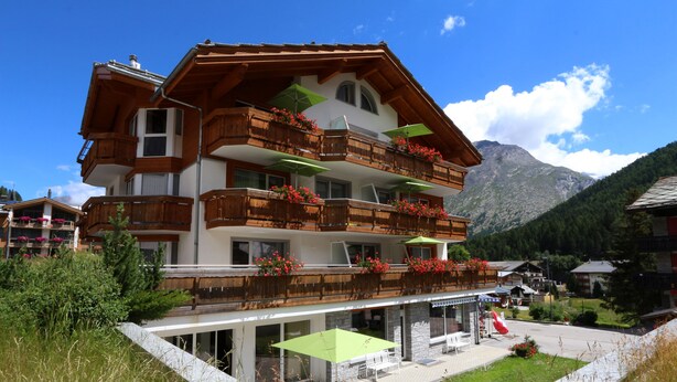 A multi-story chalet with wooden balconies, outdoor seating, and a mountain backdrop. - Apparthotel Feehof Saas-Fee (Saas-Fee)