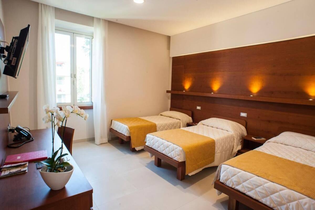 Ein Hotelzimmer mit zwei Einzelbetten, einem Holzkopfende, einem Schreibtisch mit Computer und Pflanze sowie einem Fenster mit Vorhängen.