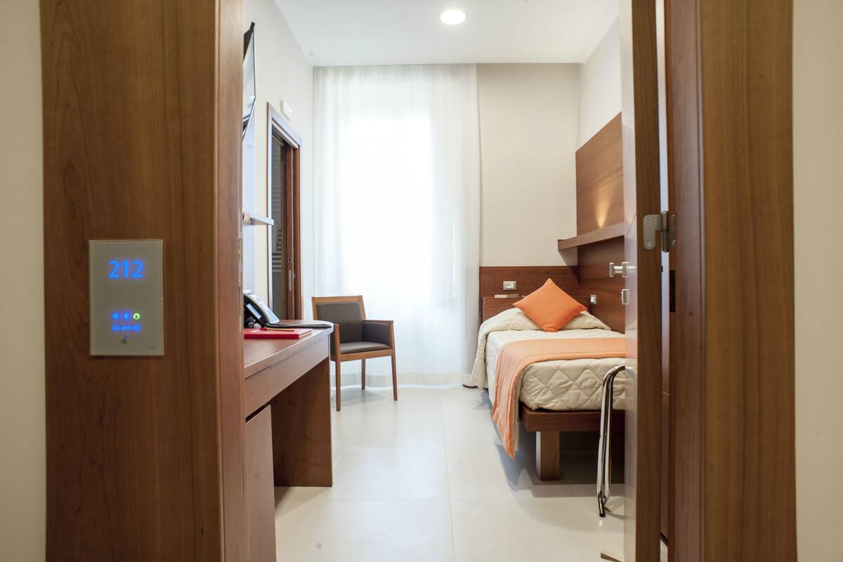 Ein Hotelzimmer mit Schreibtisch, Stuhl und einem Bett mit einem orangefarbenen Kopfkissen. An der Wand befindet sich eine Bedienfeld mit der Nummer 212.