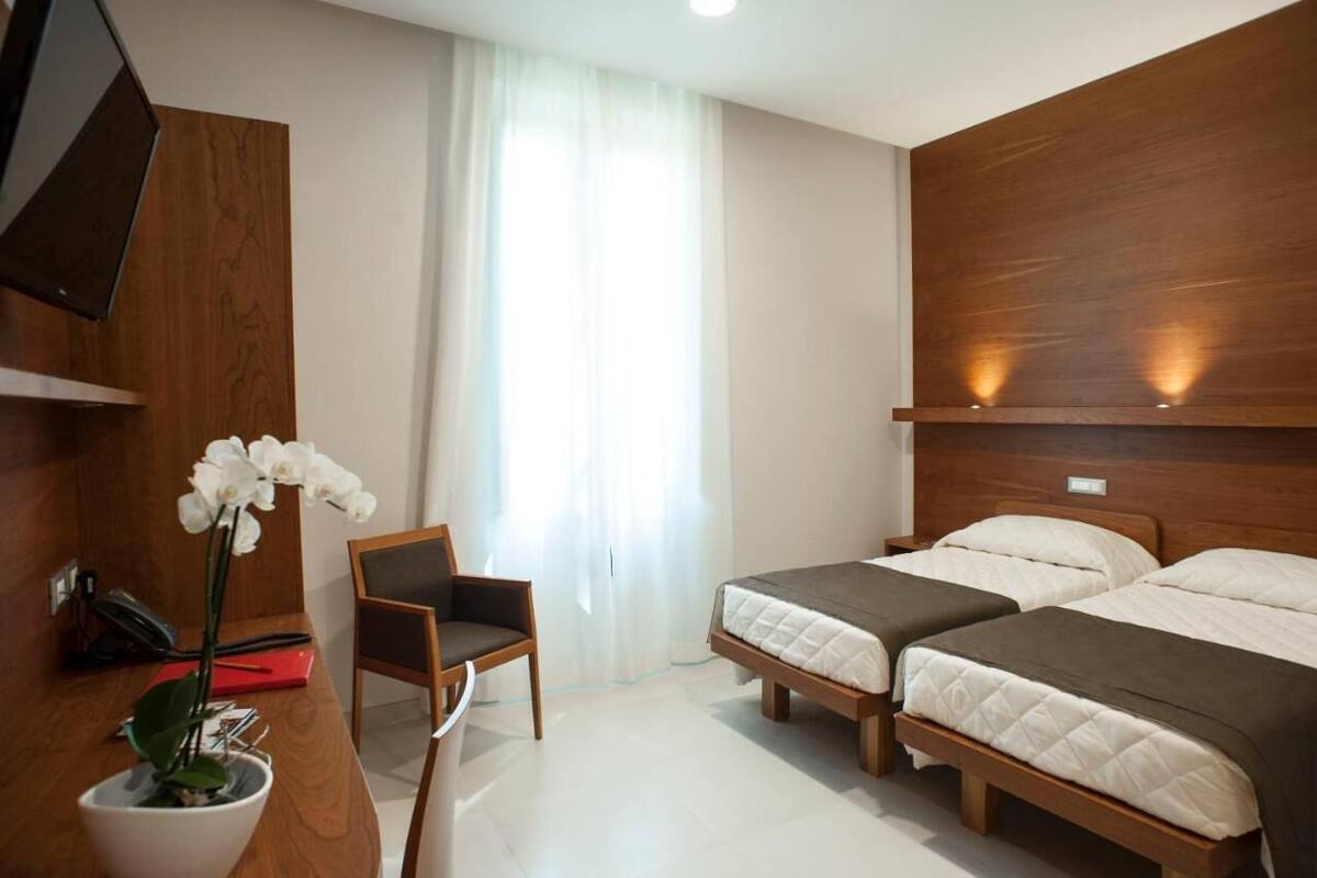 Ein Hotelzimmer mit zwei Betten, einem Schreibtisch mit Telefon, einem Sessel und einer Topf-Orchidee.