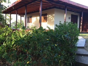 A covered outdoor area with a wooden structure, a white building with windows, and dense greenery in the foreground.