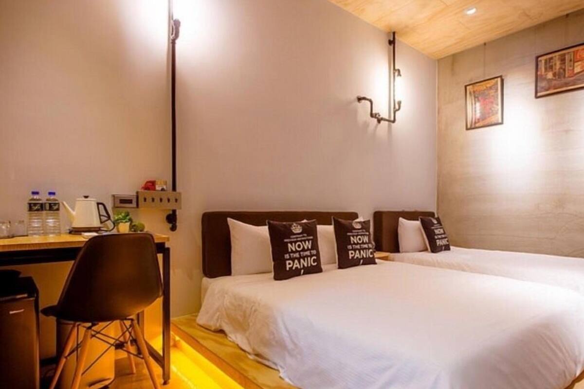 A hotel room with two beds, a desk, a chair, and a wall-mounted lamp.