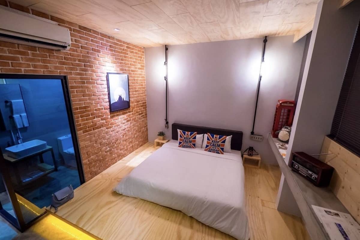 A modern bedroom with a brick wall, a bed with decorative pillows, a bedside table, a radio, and a small plant.