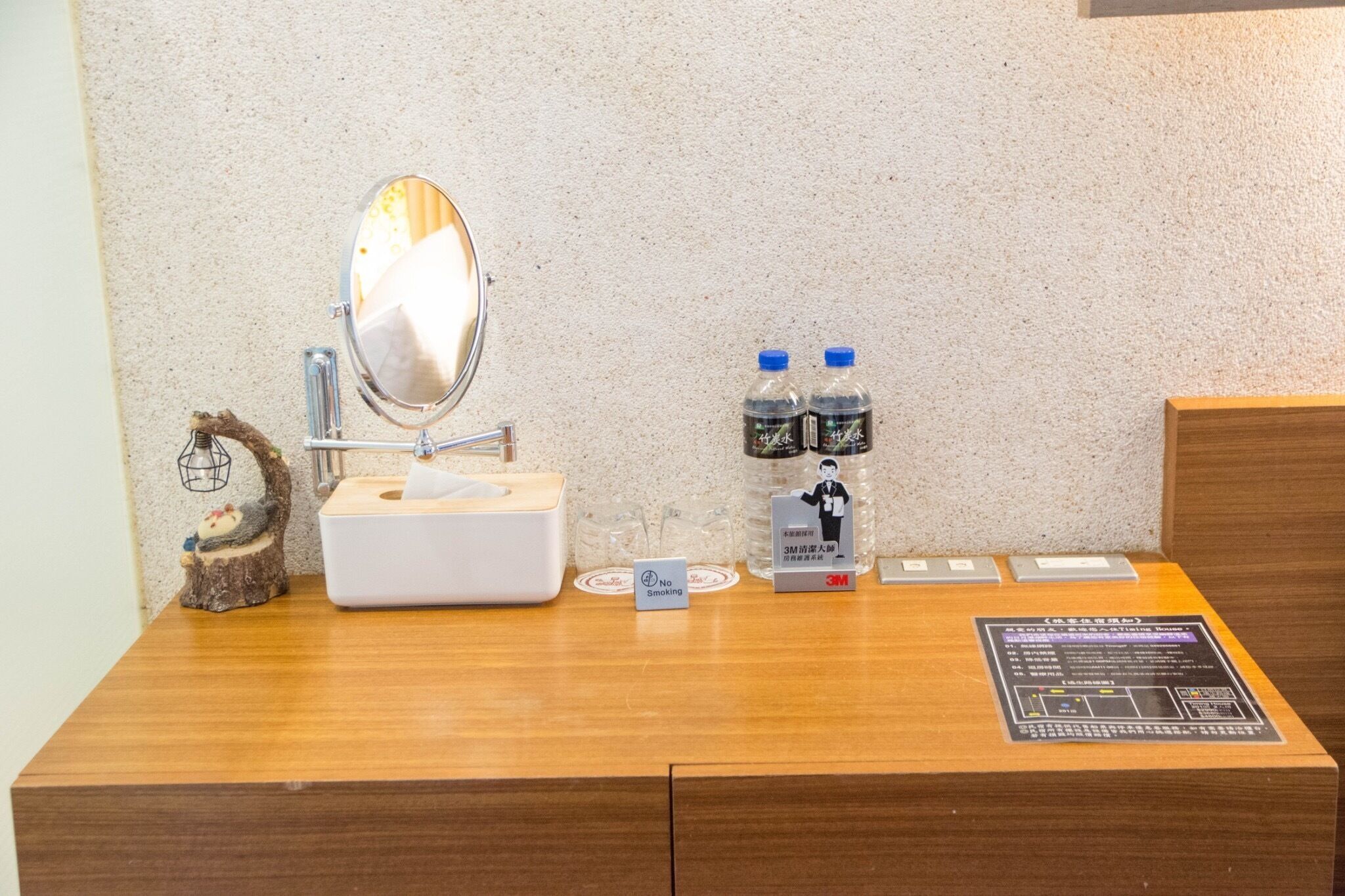 A hotel room desk with a mirror, a tissue box, two water bottles, a no-smoking sign, and a 3M sign.