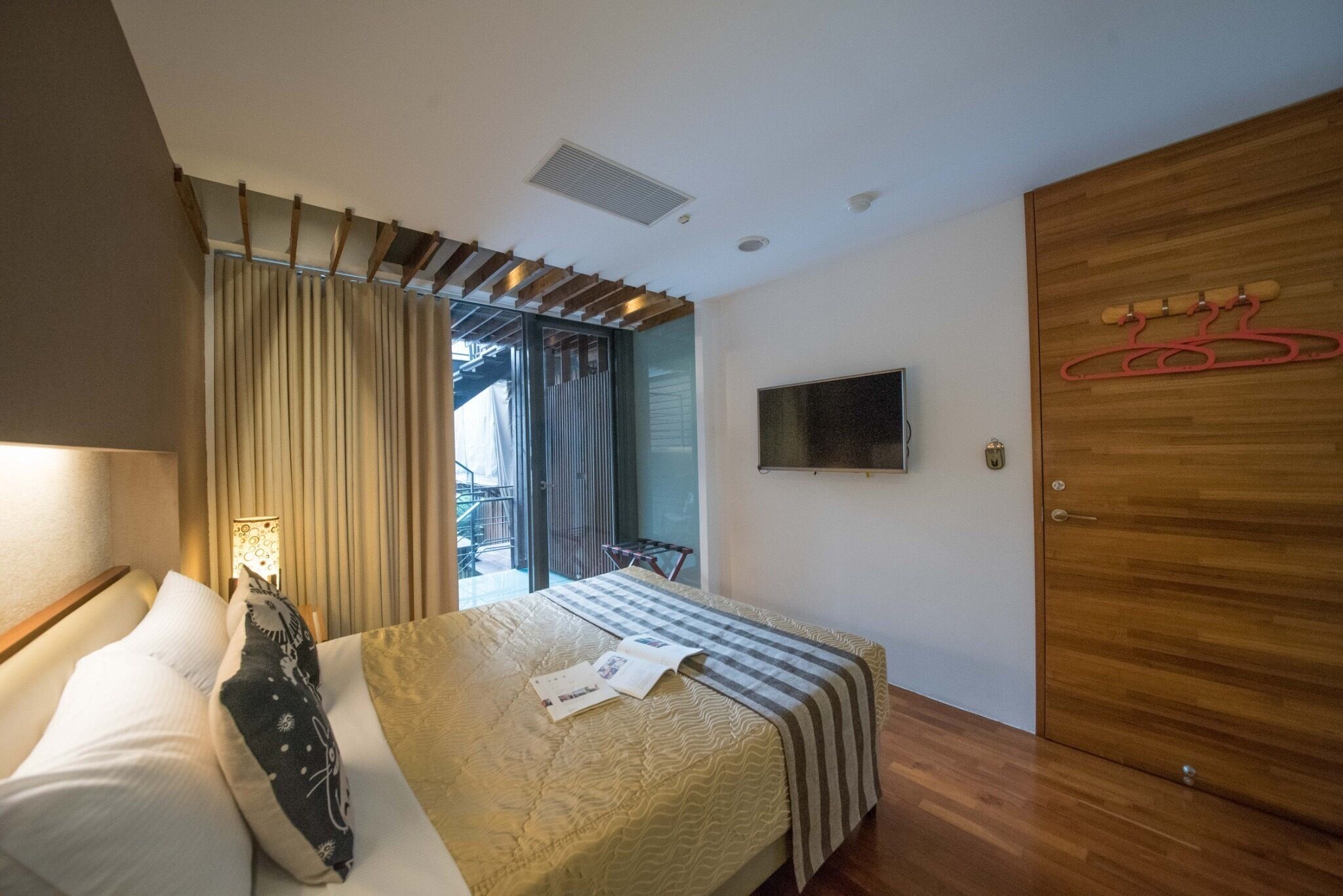 Un dormitorio con cama, televisor montado en la pared, armario de madera y puerta corrediza de vidrio que da a un balcón.