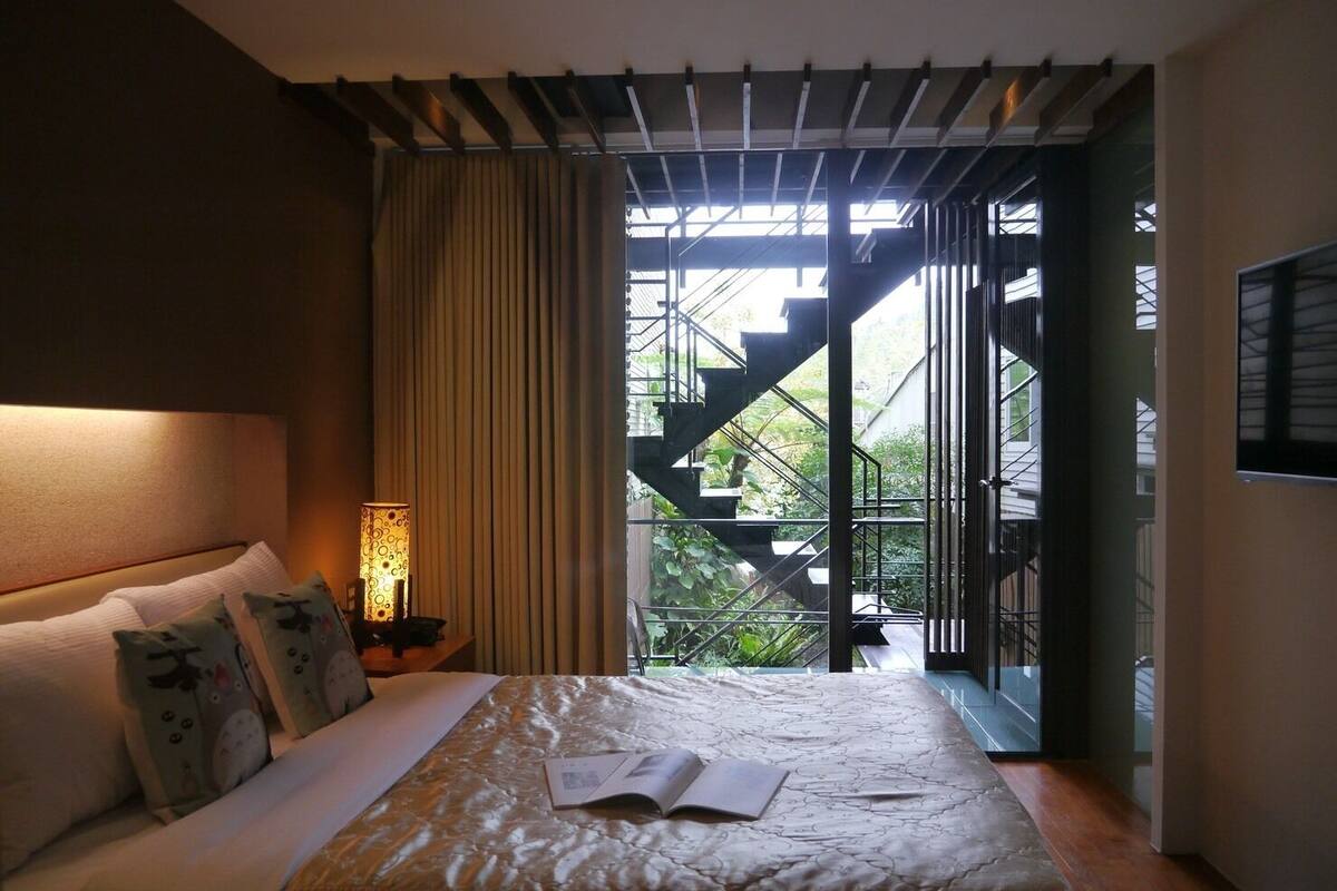 A bedroom with a bed, a bedside lamp, a window with curtains, and a view of a staircase leading outside.