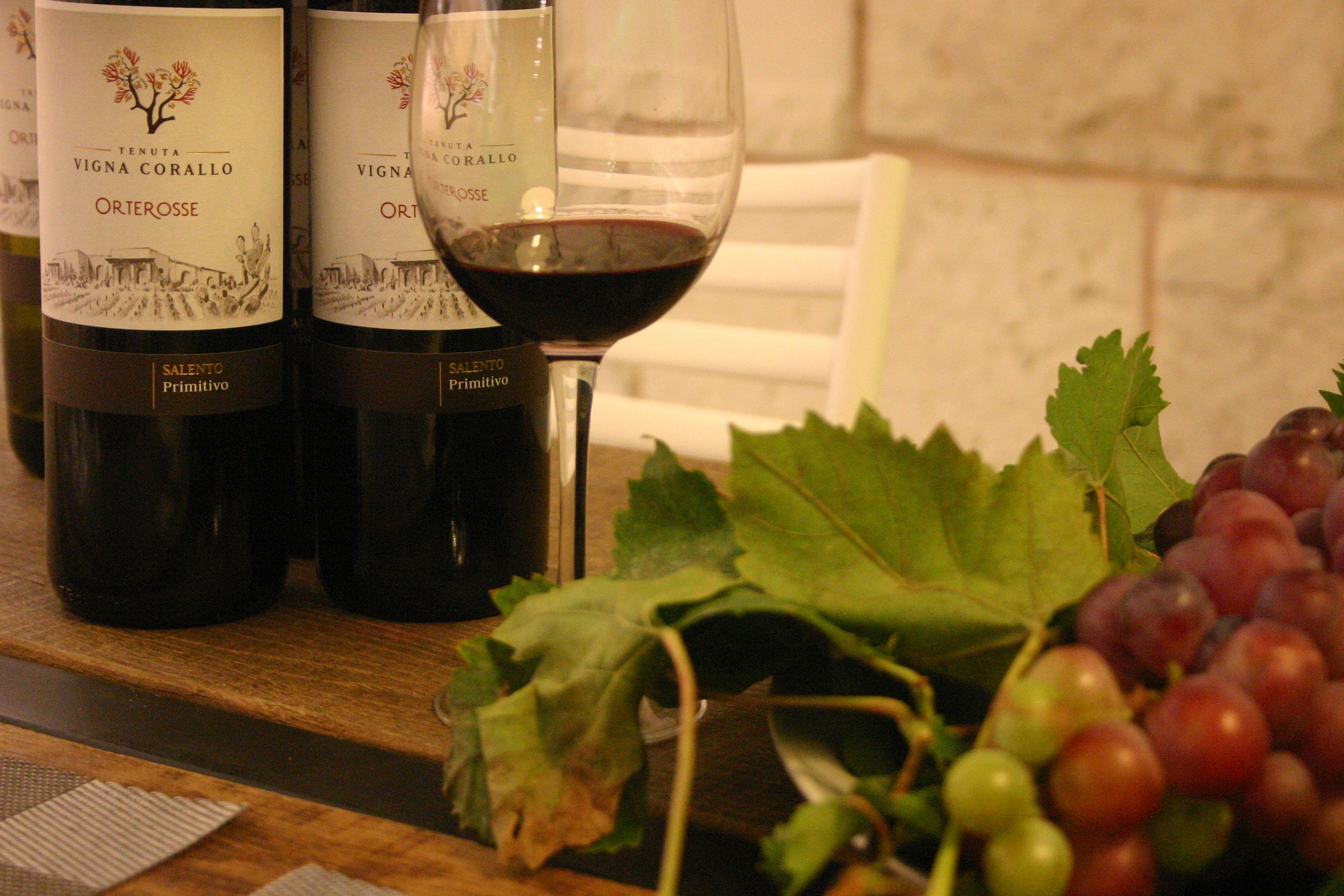 Botellas de vino Tenuta Vigna Corallo Orterosse, una copa de vino tinto y un racimo de uvas sobre una superficie de madera.
