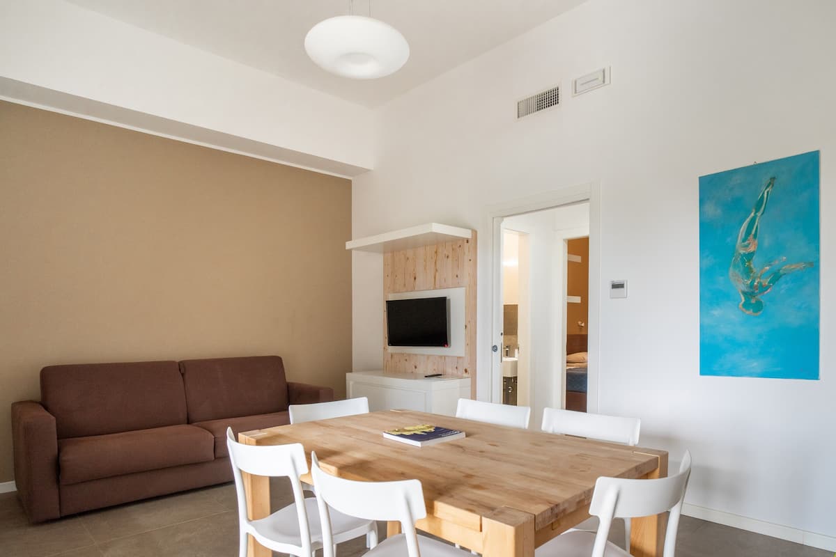 A modern living room with a brown sofa, a wooden dining table with white chairs, a flat-screen TV mounted on a wooden panel, and a blue painting on the wall.