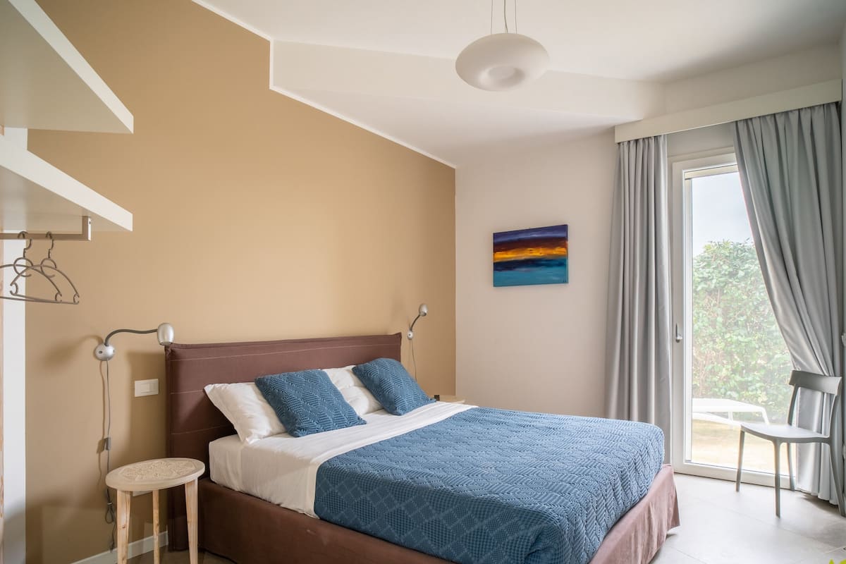 A bedroom with a bed, a bedside table, a chair, a painting, and a window with curtains.