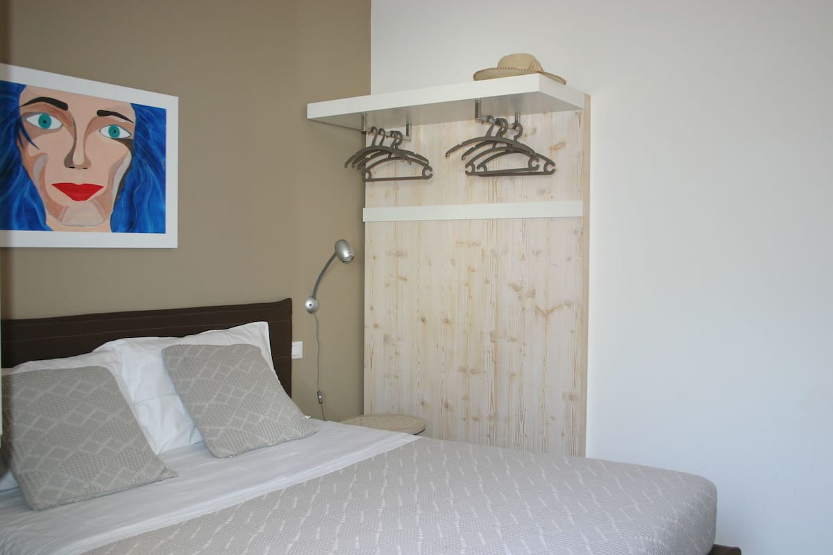 A bedroom with a bed, pillows, a lamp, a wooden wardrobe, and a hat on the shelf.