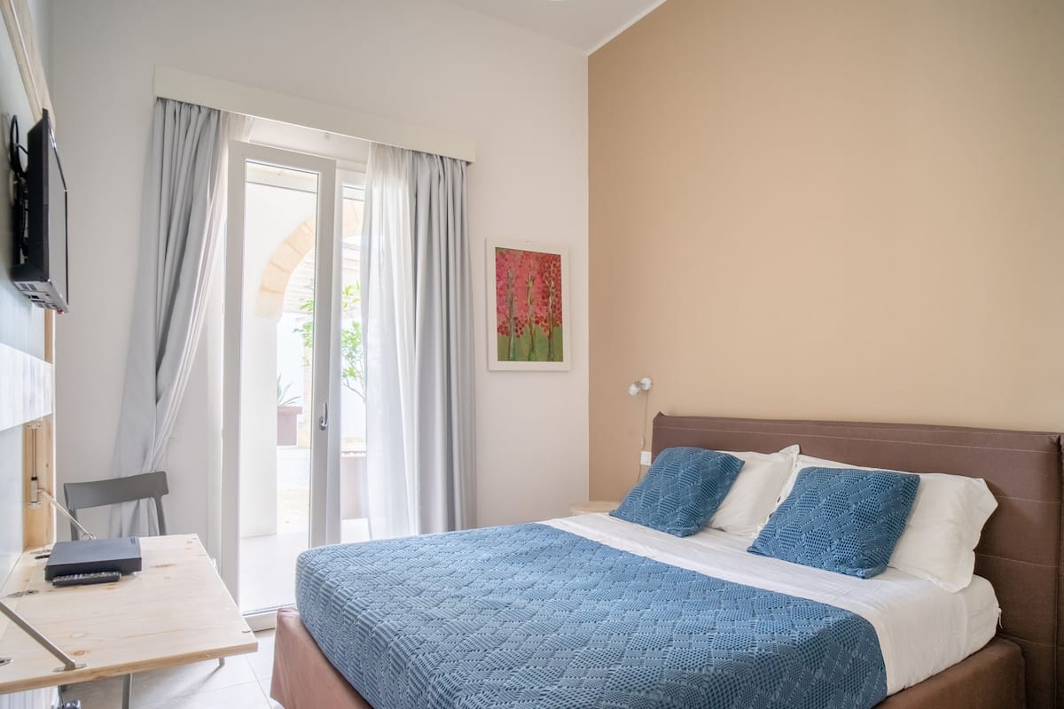 A bedroom with a bed, a desk, a chair, a TV, and a window with curtains.