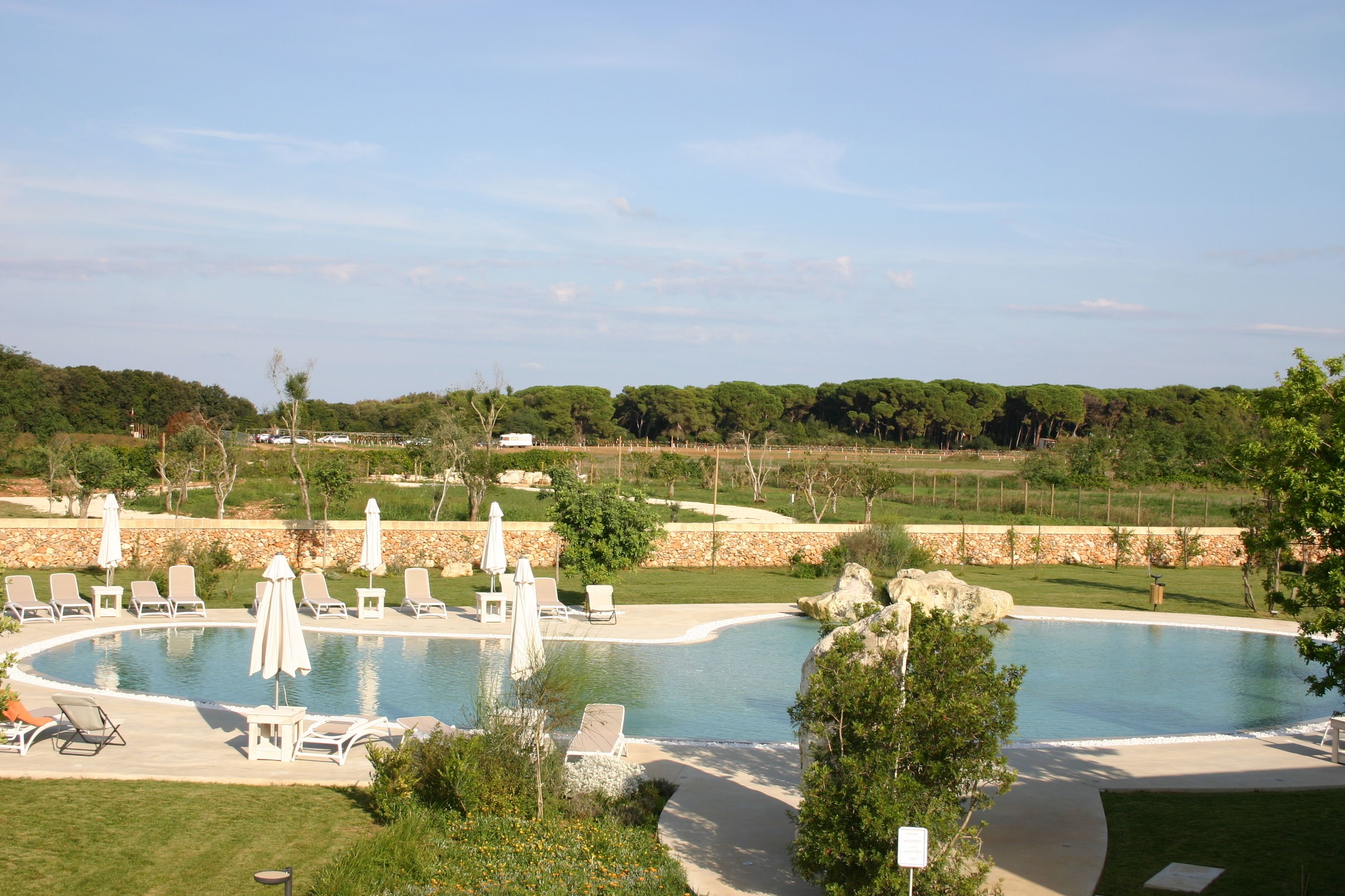 Zona de piscina con sillas de descanso y sombrillas, rodeada de vegetación y un muro de piedra.