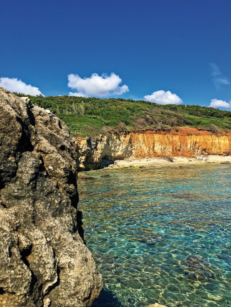 A coastal landscape with clear water, rocky formations, and a green hillside.