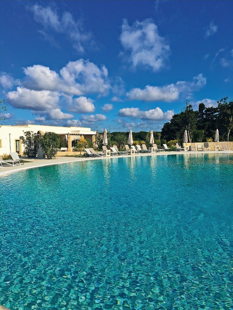 Une piscine d’un bleu éclatant, bordée de chaises longues et d’ombrelles, entourée d’un bâtiment à la façade en pierre, sous un ciel dégagé.