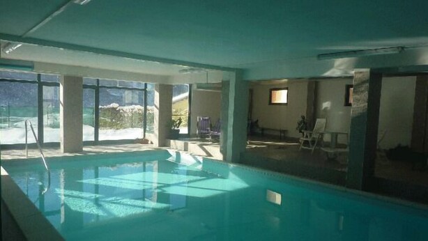 An indoor swimming pool with clear glass walls, a view of the outdoors, and a seating area with chairs and a table. - Hotel Campo Felice (Lucoli)