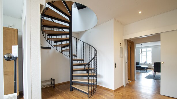 Un interior moderno con una escalera espiral, suelo de madera y vista a un salón comedor con mesa y sillas.