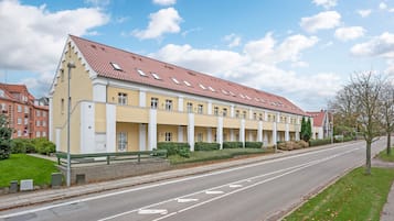 Ein langgestrecktes, gelbes Gebäude mit rotem Ziegeldach und zahlreichen Fenstern, an einer breiten Straße mit Bürgersteig und Grünflächen gelegen.