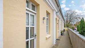 Un pasillo estrecho con fachada color beige, ventanas de marco blanco y una pequeña zona exterior con plantas en macetas.