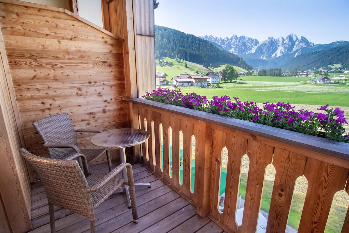 A wooden deck with wicker chairs and a small table, overlooking a green landscape and mountains.