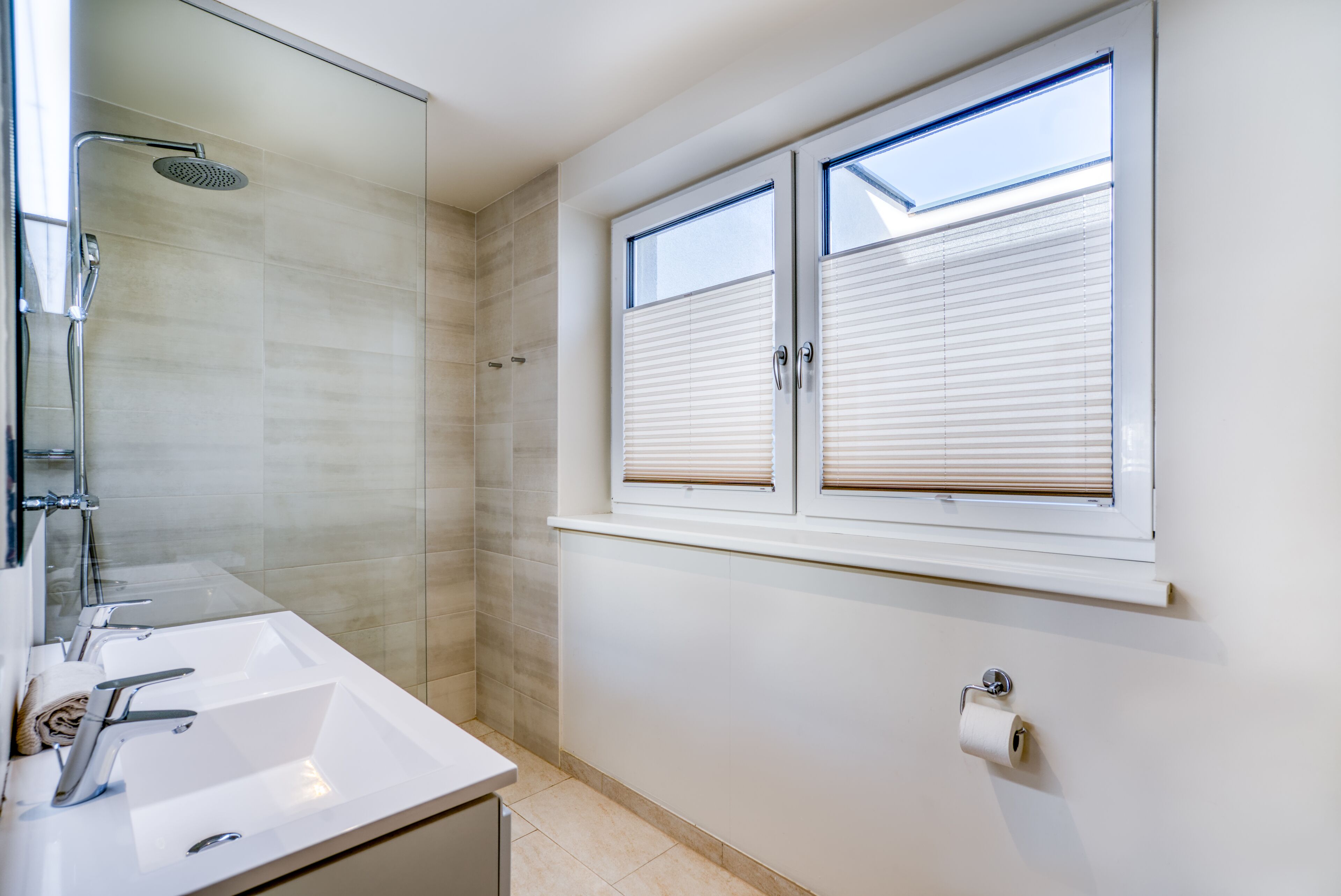 A modern bathroom with a shower, two sinks, a toilet, and a window with blinds.