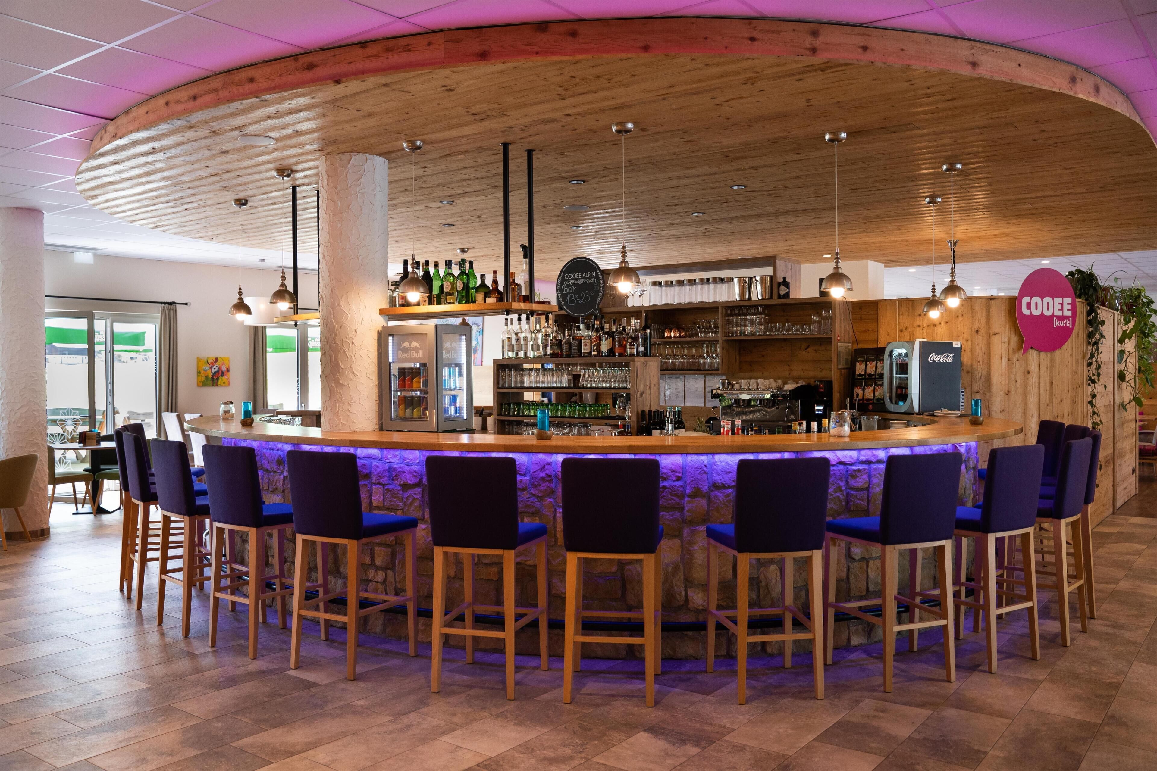 Zona de bar con mostrador de madera, varios taburetes y una gran variedad de botellas y vasos.