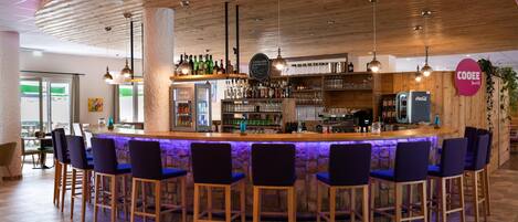 Zona de bar con mostrador de madera, varios taburetes y una gran variedad de botellas y vasos.