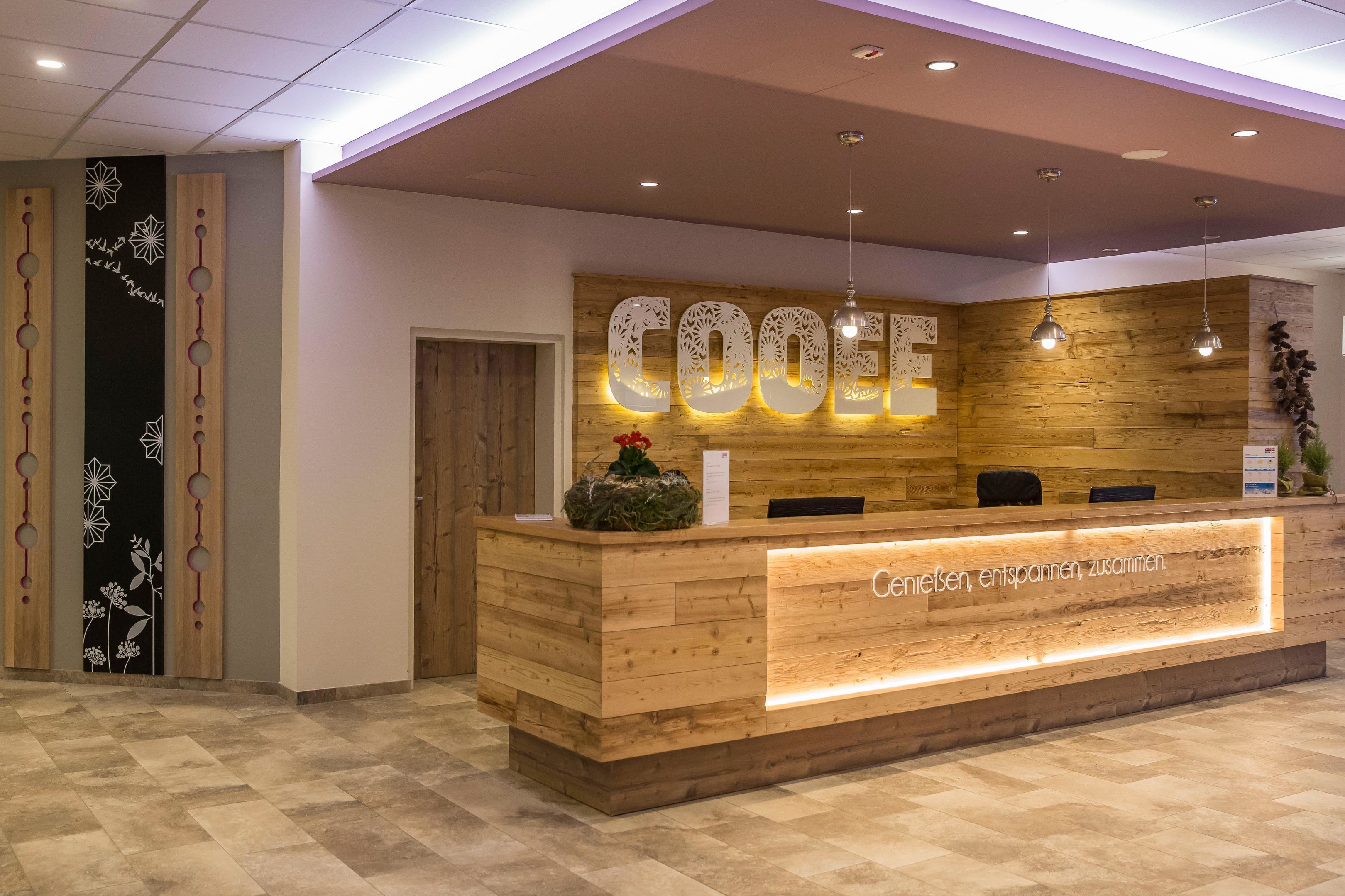 A reception desk with a wooden finish, a wall-mounted sign with the word 'COOEE,' and a reception area with a wooden wall and hanging pendant lights.