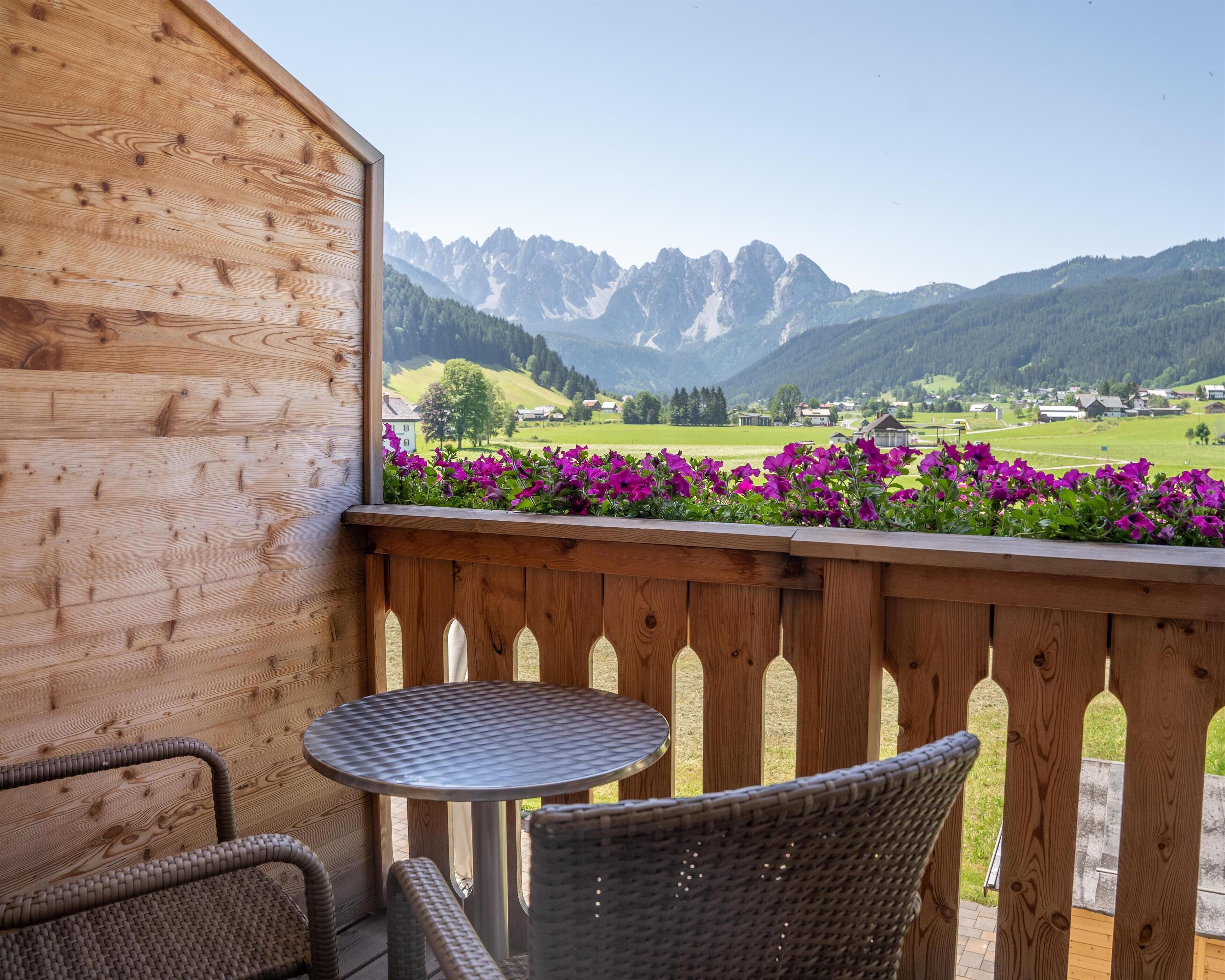 Ein Holzbalkon mit Tisch und Stühlen, der auf eine Berglandschaft mit violetten Blumen im Vordergrund blickt.