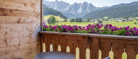 Ein Holzbalkon mit Tisch und Stühlen, der auf eine Berglandschaft mit violetten Blumen im Vordergrund blickt.
