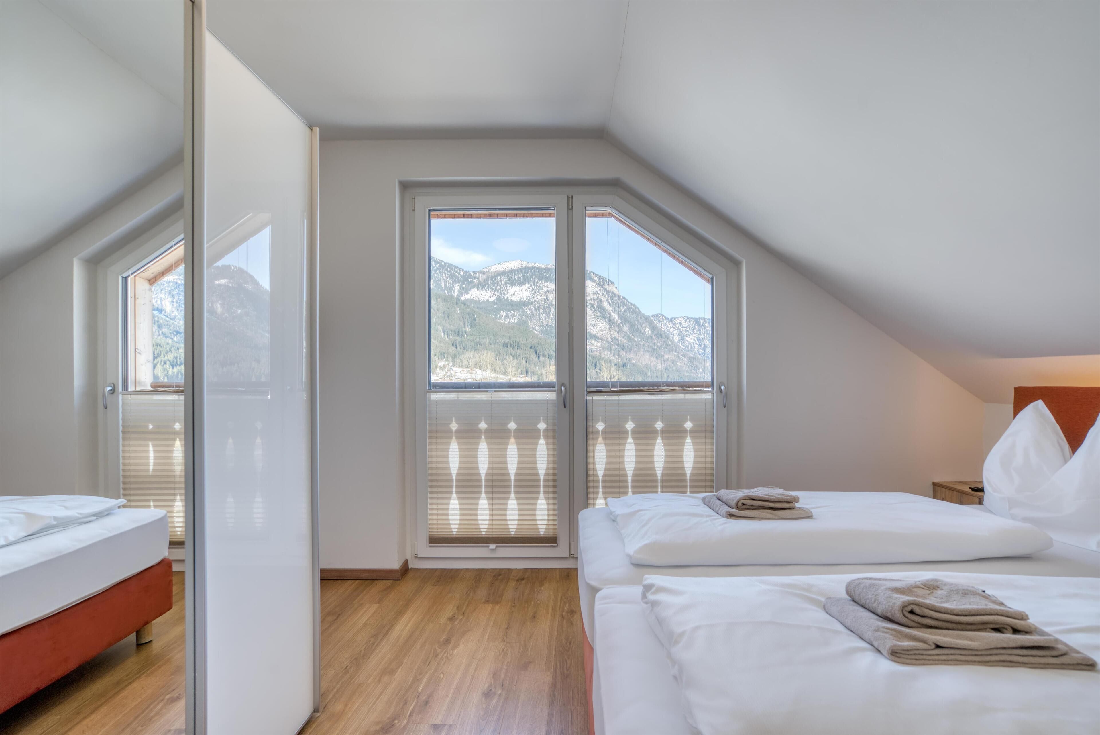 A bedroom with a wooden floor, two beds, a large window with a view of mountains, and a sliding door.