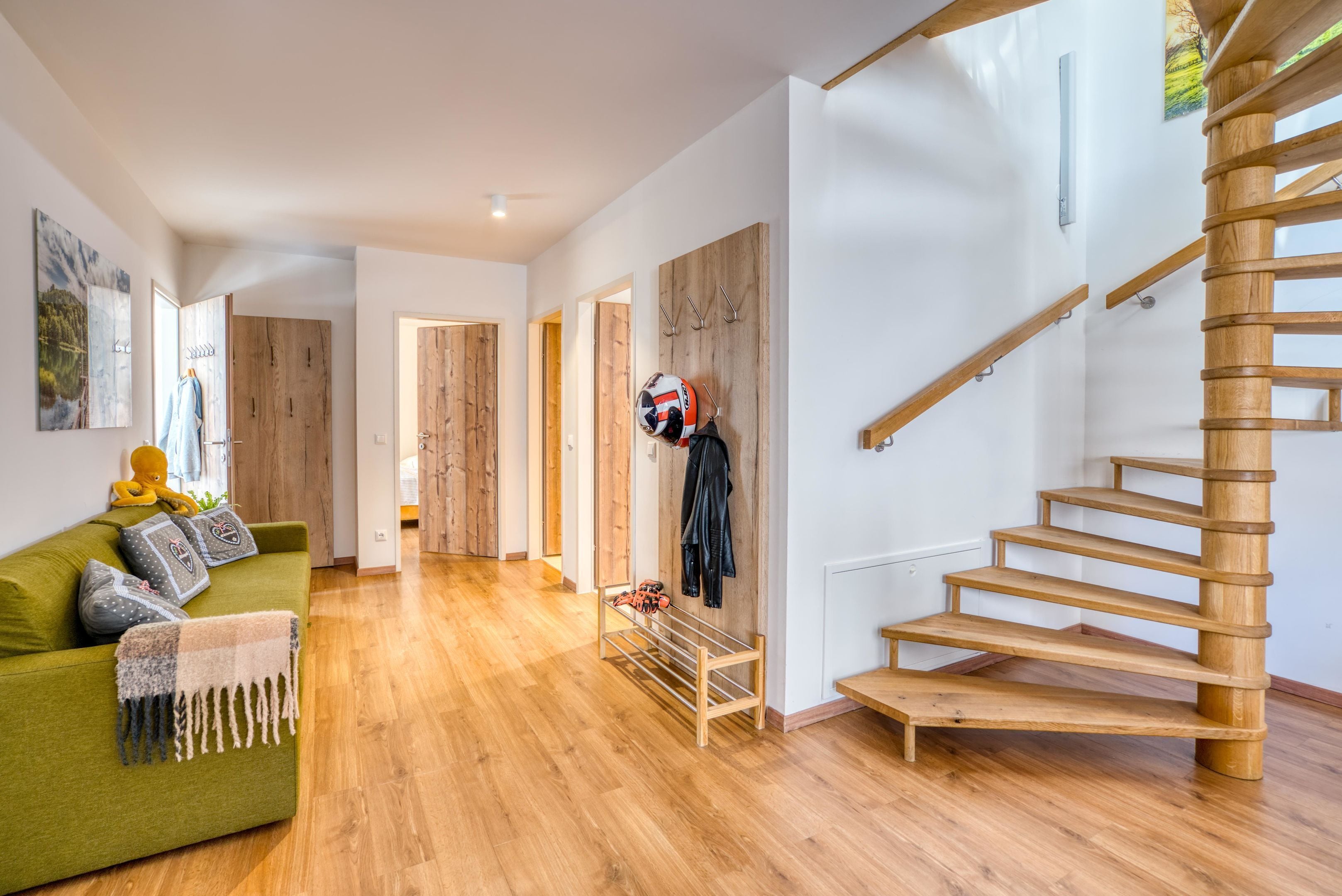 A modern interior with a wooden staircase, a green sofa with cushions, and a wall-mounted rack with a helmet and clothes.