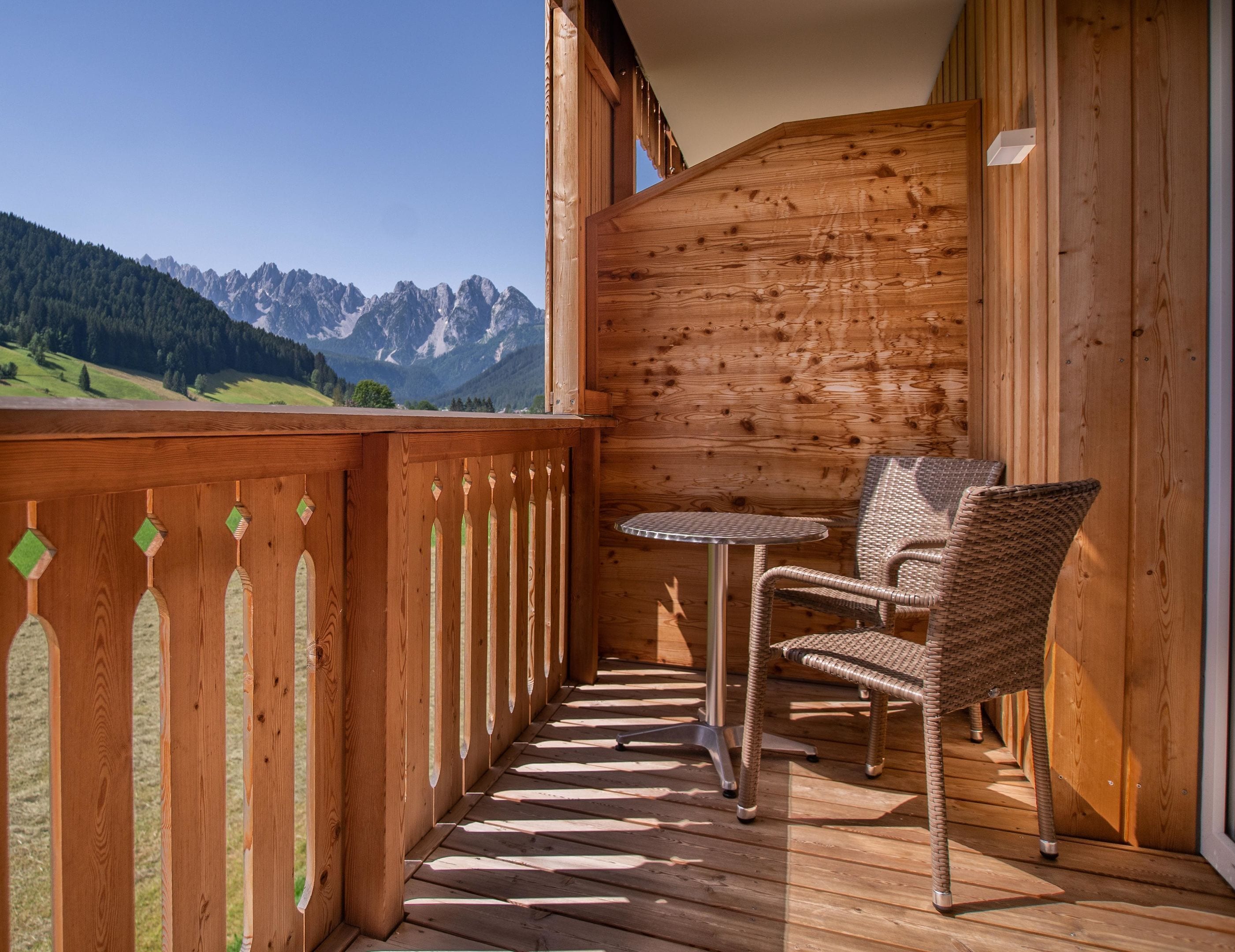 A wooden balcony with a view of mountains, a table, and two wicker chairs.