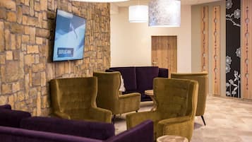 Un hall d’entrée avec un mur en pierre, un écran de télévision et des espaces de détente dotés de fauteuils violets et dorés.