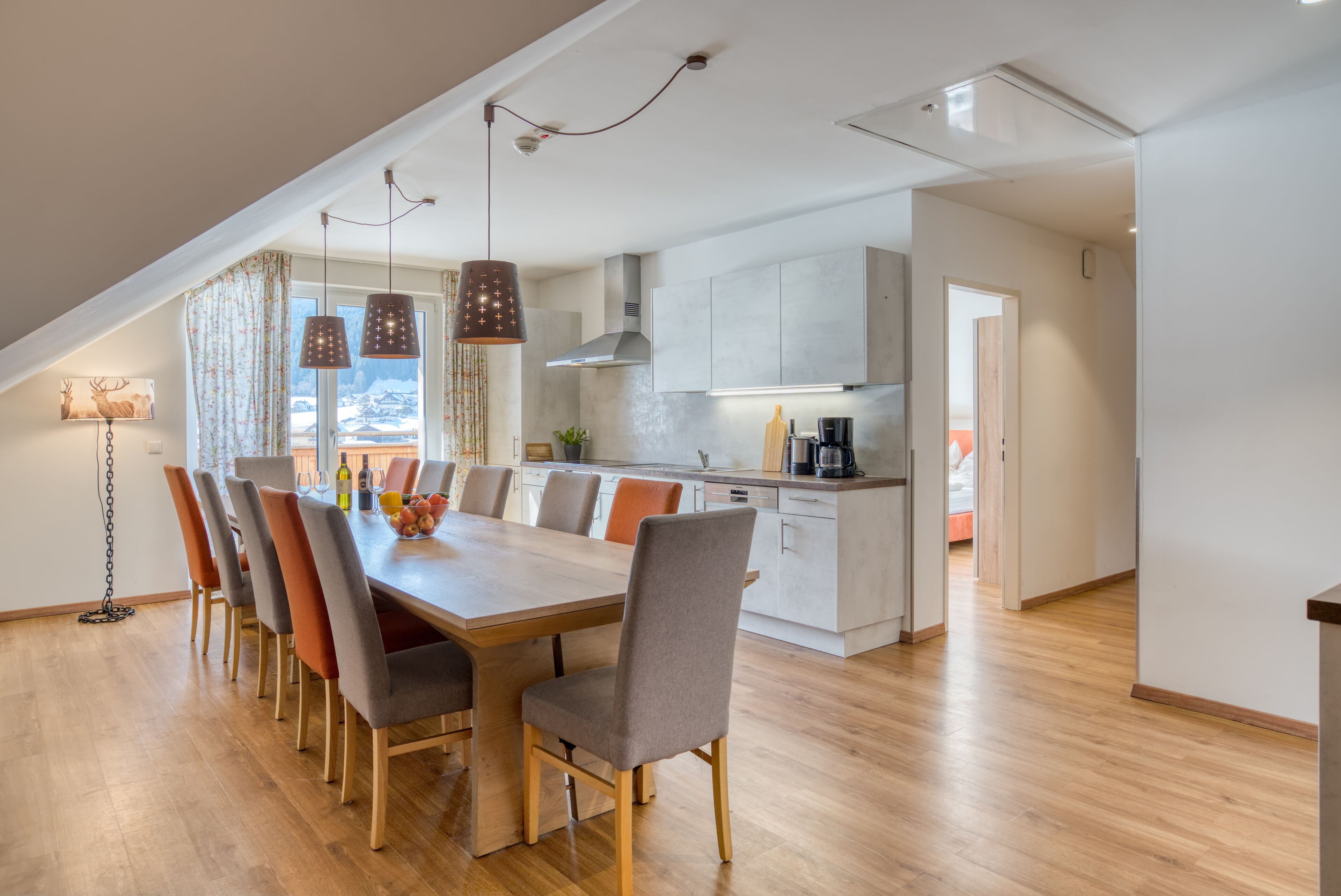 Een moderne keuken met een houten eettafel, gestoffeerde grijze stoelen, hanglampen en uitzicht op een besneeerd landschap.