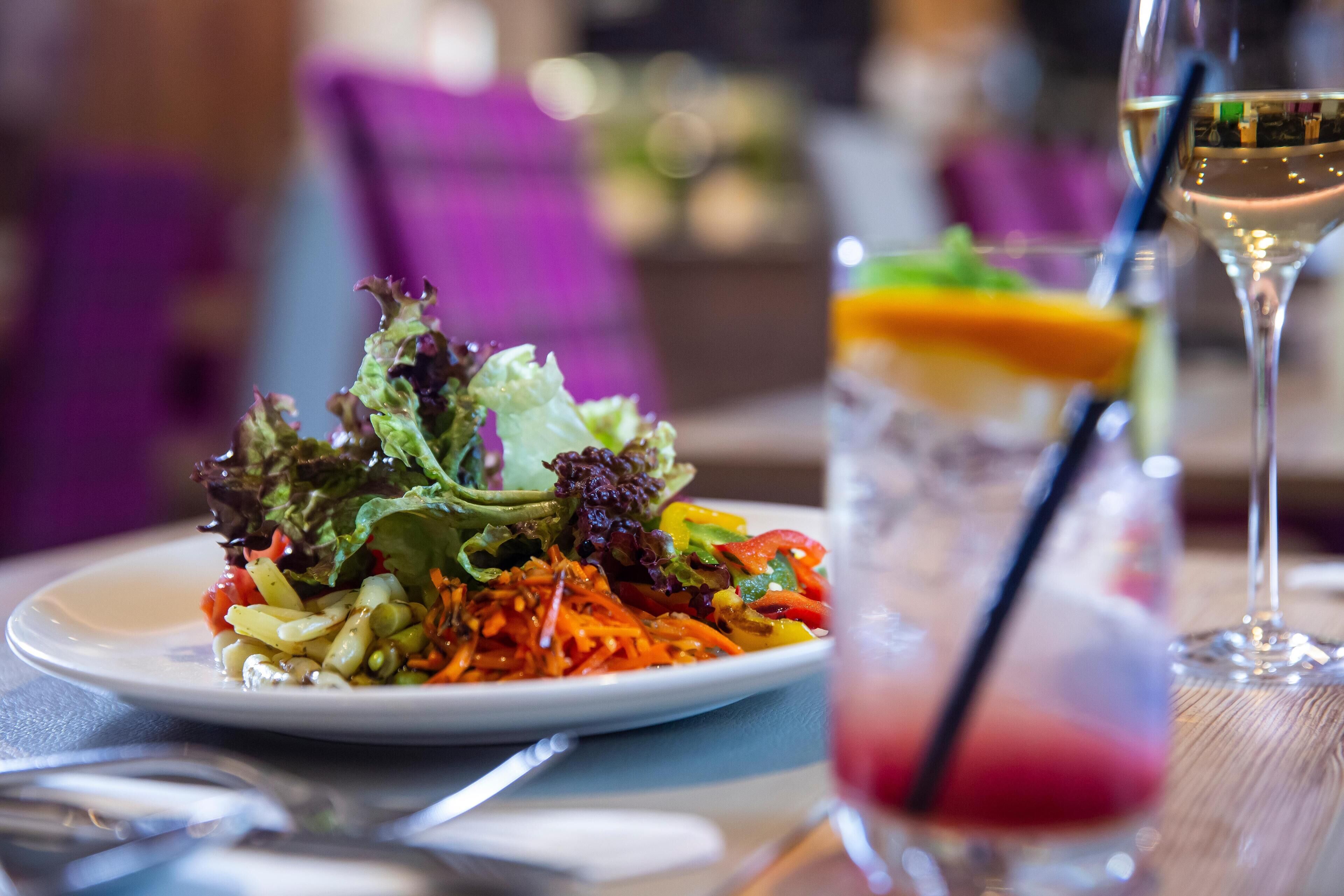 A plate of mixed greens and vegetables, a glass of white wine, and a cocktail with a straw.