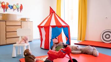 A playroom with a red and white striped tent, a small white chair, a red toy car, and a teddy bear.