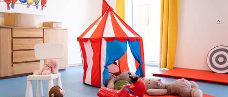 Una sala de juegos con una carpa a rayas rojas y blancas, una pequeña silla blanca, un coche de juguete rojo y un osito de peluche.