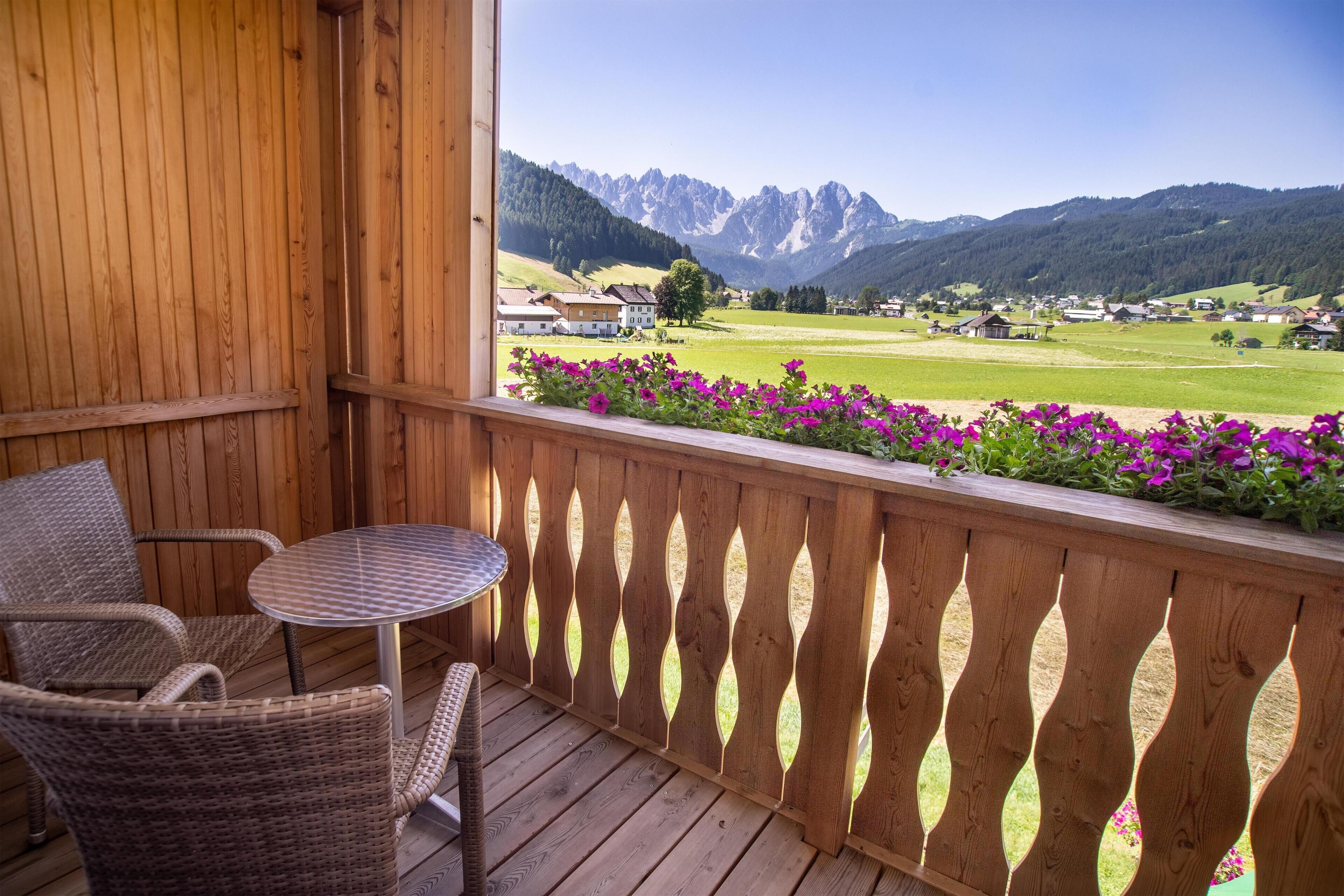 A wooden balcony with wicker chairs and a small table, overlooking a green valley and mountains.